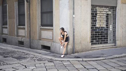 Slim female runner in activewear standing on corner of street and tying shoelaces on sneakers while leaning on stone wall of building and looking away