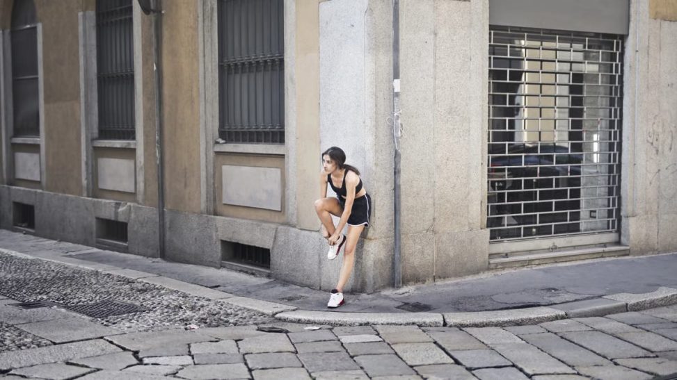 Slim female runner in activewear standing on corner of street and tying shoelaces on sneakers while leaning on stone wall of building and looking away